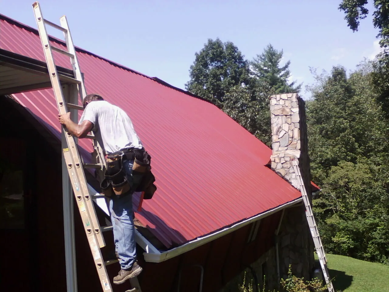 Roofer on a ladder beside a red metal roof and stone chimney — Soffit & Fascia Repair by Roofing Brothers in Goodfield